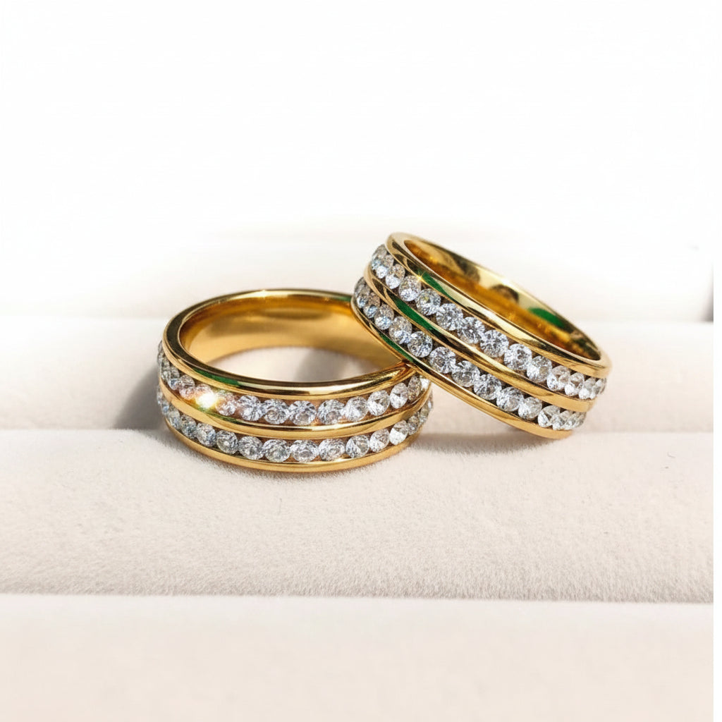 Two gold rings with rows of clear pavé stones, displayed on a beige jewelry box.