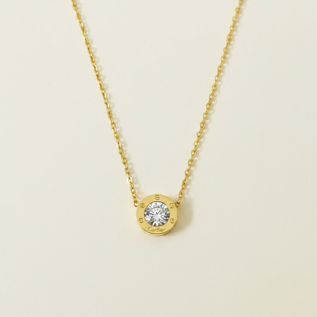 A gold chain necklace with a round gold pendant featuring a clear central gemstone and a small gold logo at the bottom, displayed on a dark gray background.
