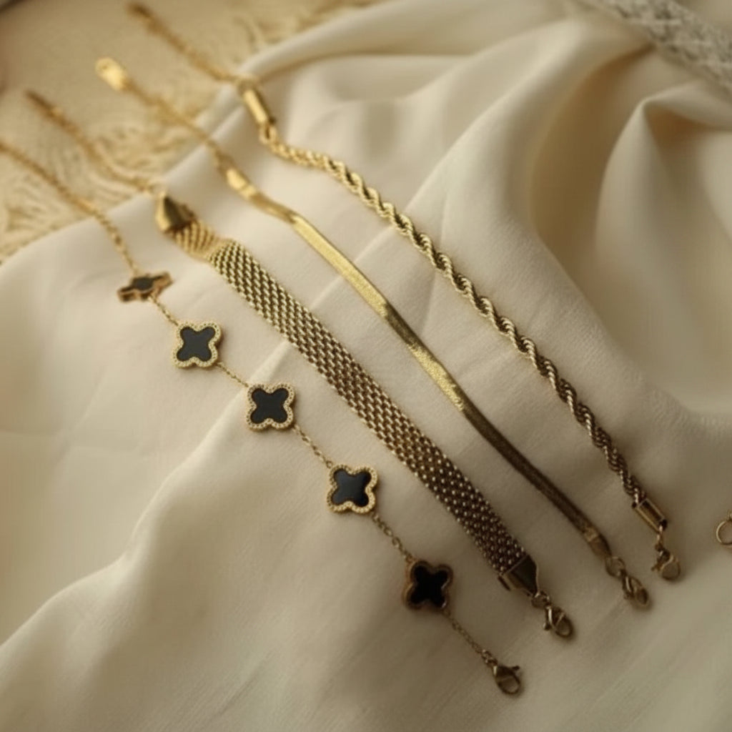 Four gold-toned chain bracelets with different textures and designs, including three with black inlay clover motifs, arranged on draped cream-colored fabric.