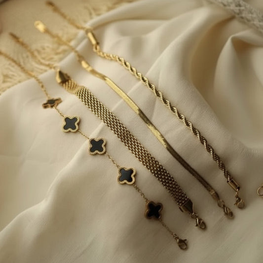 Four gold-toned chain bracelets with different textures and designs, including three with black inlay clover motifs, arranged on draped cream-colored fabric.