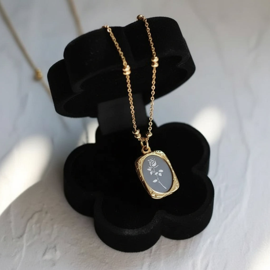 A gold pendant necklace with a rectangular gray stone featuring white floral engraving, displayed on a black flower-shaped jewelry stand against a textured white background.