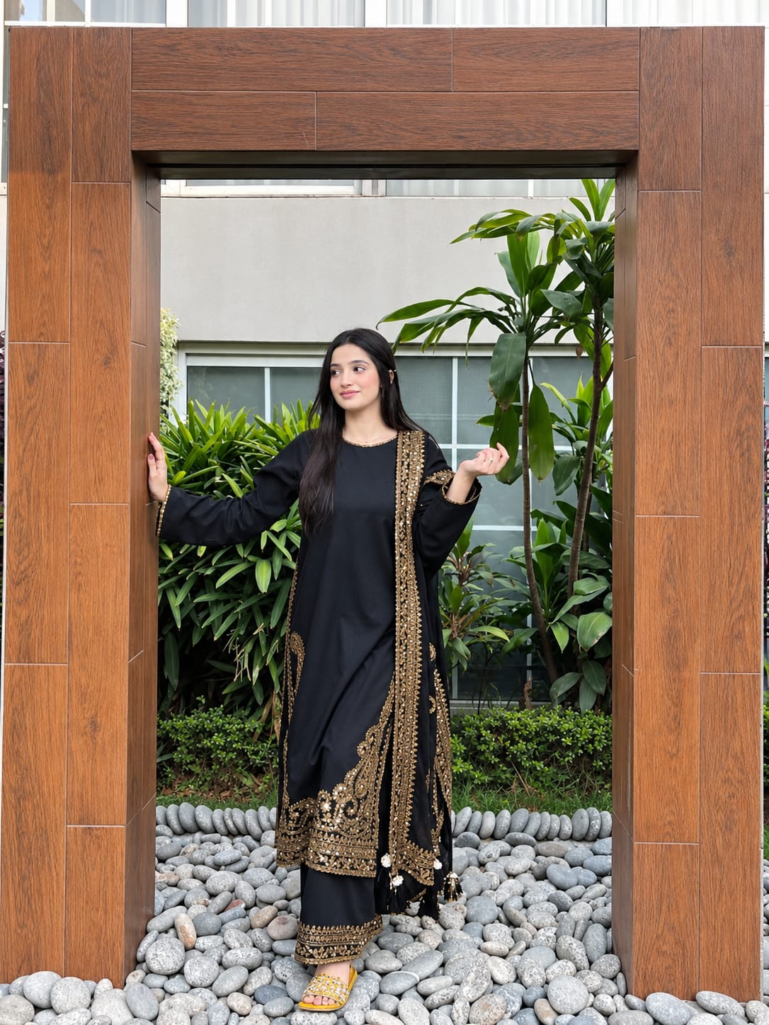 A woman wearing a black traditional outfit with ornate gold embroidery stands inside a large wooden frame, with green plants and a stone pebble ground behind her.