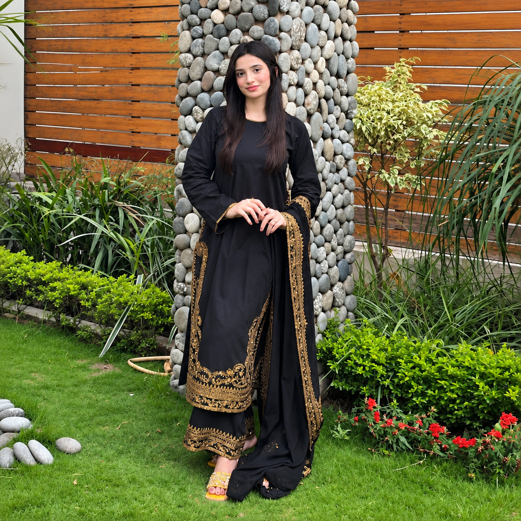 A woman stands outdoors on a lawn in front of a stone pillar and wooden slat wall, wearing a black traditional outfit with ornate gold embroidery and a matching dupatta.