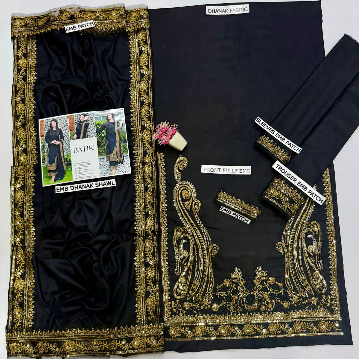 Black and gold embroidered fabric set laid out on a white background, including a large embroidered border piece, a matching black fabric panel, folded fabric pieces, and a small floral prop. Visible text labels include 'EMB DHANAK SHAWL', 'DHANAK PULLI', and 'DHANAK BANDA'.