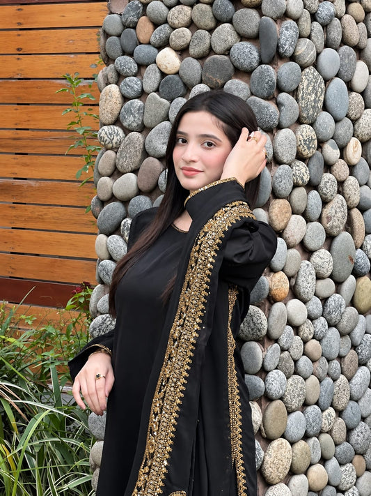 A woman wearing a black traditional outfit with ornate gold embroidery along the border, posing in front of a stone wall and wooden slat background.