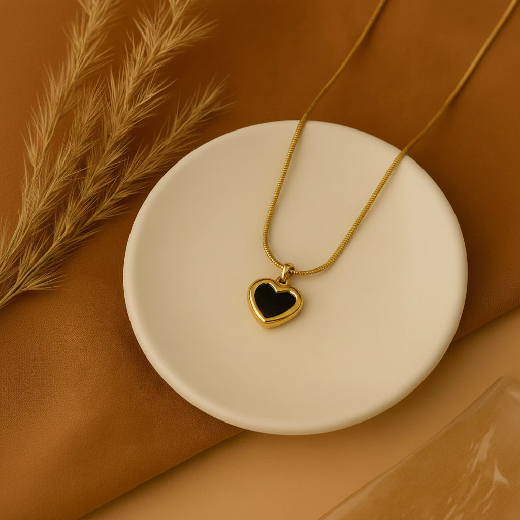 A gold heart pendant necklace with a black center, displayed on a round white ceramic dish over a warm brown textured background with dried grass.