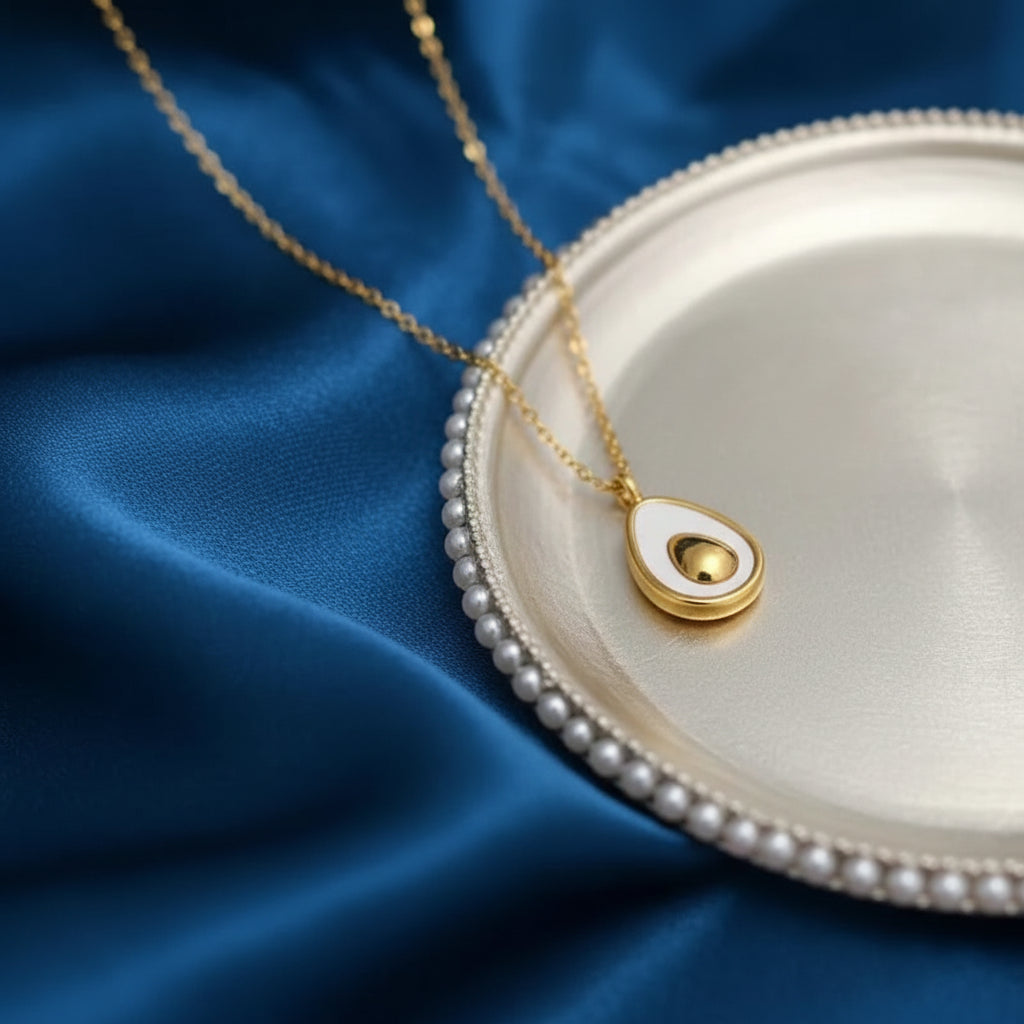 Gold chain necklace with a teardrop-shaped pendant featuring a white inlay and a gold center, displayed on a silver tray against a blue satin background.