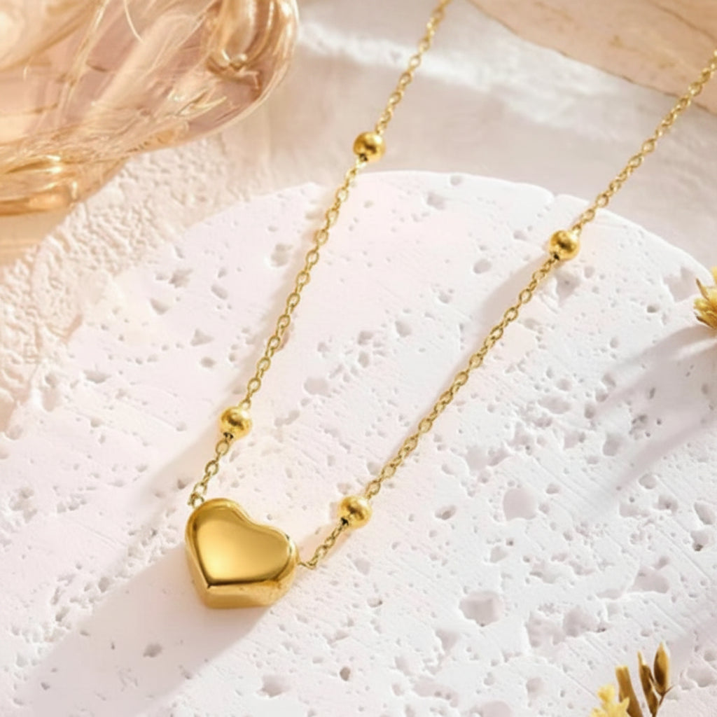 A gold heart pendant necklace with small gold bead accents on a delicate chain, displayed on a textured white surface.