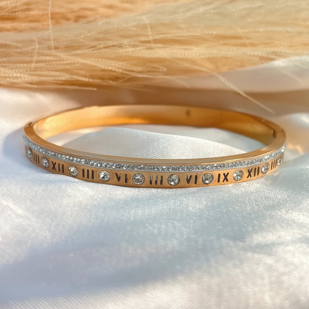 A gold bangle bracelet with Roman numerals and clear rhinestones, displayed on a white satin fabric with a beige textured background.
