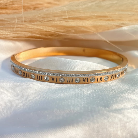 A gold bangle bracelet with Roman numerals and clear rhinestones, displayed on a white satin fabric with a beige textured background.