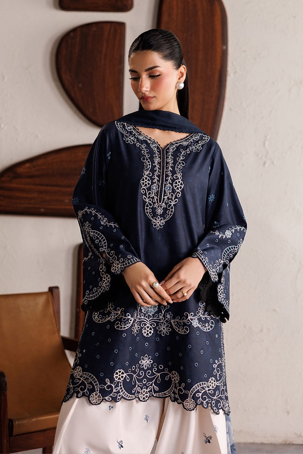 Woman wearing a navy blue embroidered traditional outfit with white pants, standing against a neutral wall.