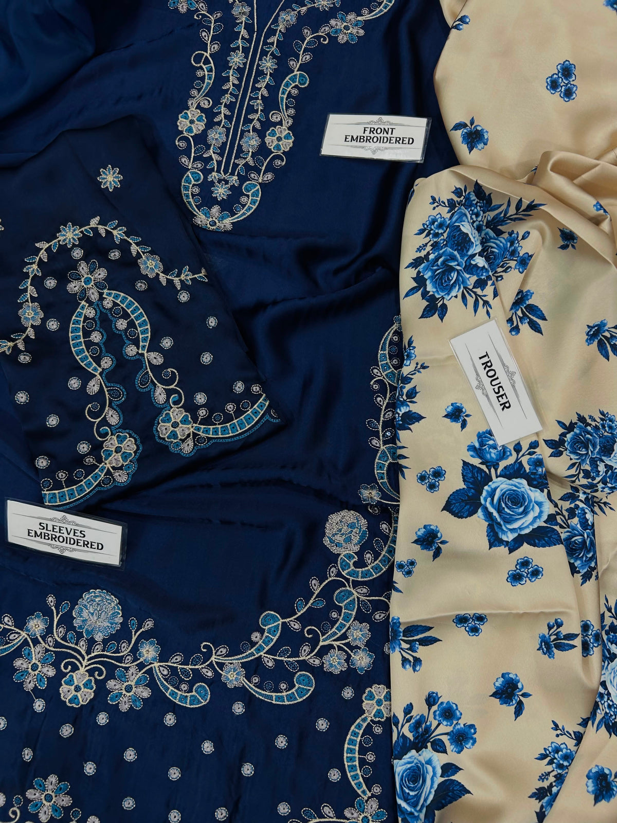A folded view of a traditional embroidered garment showing the front and sleeve embroidery in navy blue, alongside a cream-colored trouser leg with a blue floral print. White labels are visible, including one that reads 'TROUSSE'.