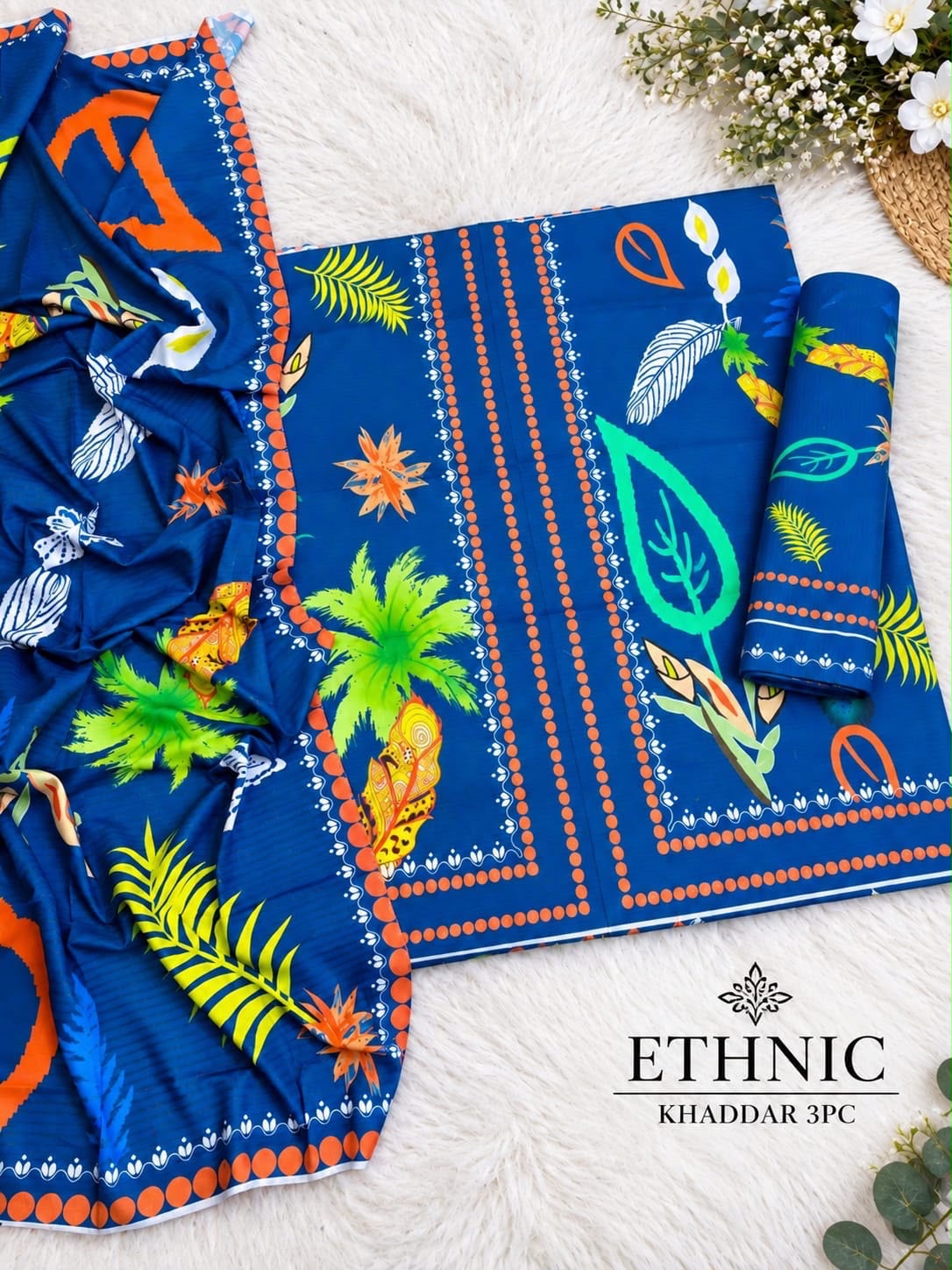 A blue printed fabric set laid out on a white faux-fur surface, including a flat sheet, a rolled fabric piece, and a matching dupatta. The print features tropical leaves, palm trees, birds, and orange geometric border details.