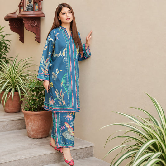 A woman models a blue printed kurta and trouser set with colorful floral and bird motifs, standing on stone steps in front of a beige wall with potted plants.