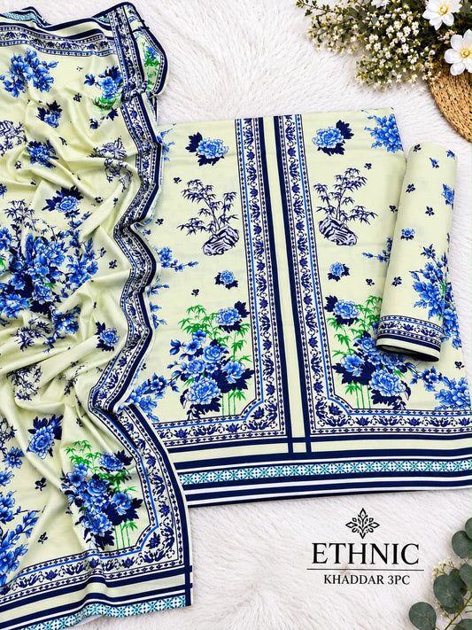 A folded and draped piece of floral printed fabric laid out on a white faux-fur surface, with matching border details visible. The fabric has a pale yellow-green base with blue and green floral motifs and navy decorative borders.