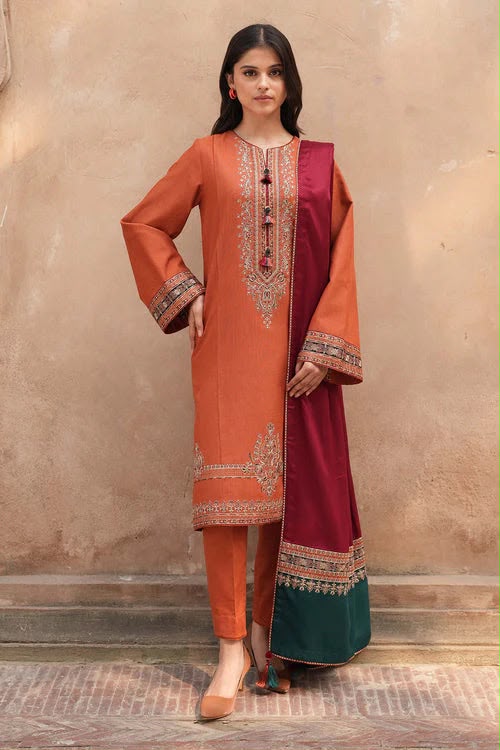 A woman models a traditional three-piece salwar kameez suit. The kurta is burnt orange with intricate gold and silver embroidery on the chest and cuffs, paired with matching orange trousers and a deep maroon dupatta featuring a green border and ornate gold embroidery.