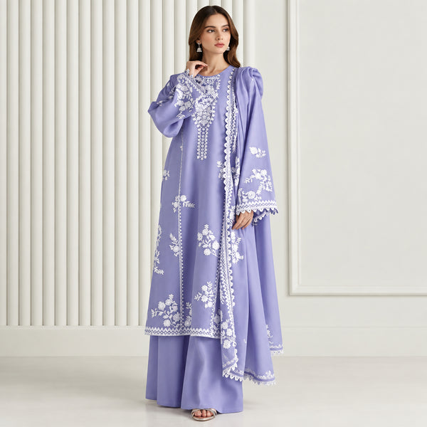 A woman models a lavender purple traditional outfit with white floral embroidery, featuring a long tunic and matching wide-leg pants, set against a neutral beige paneled background.