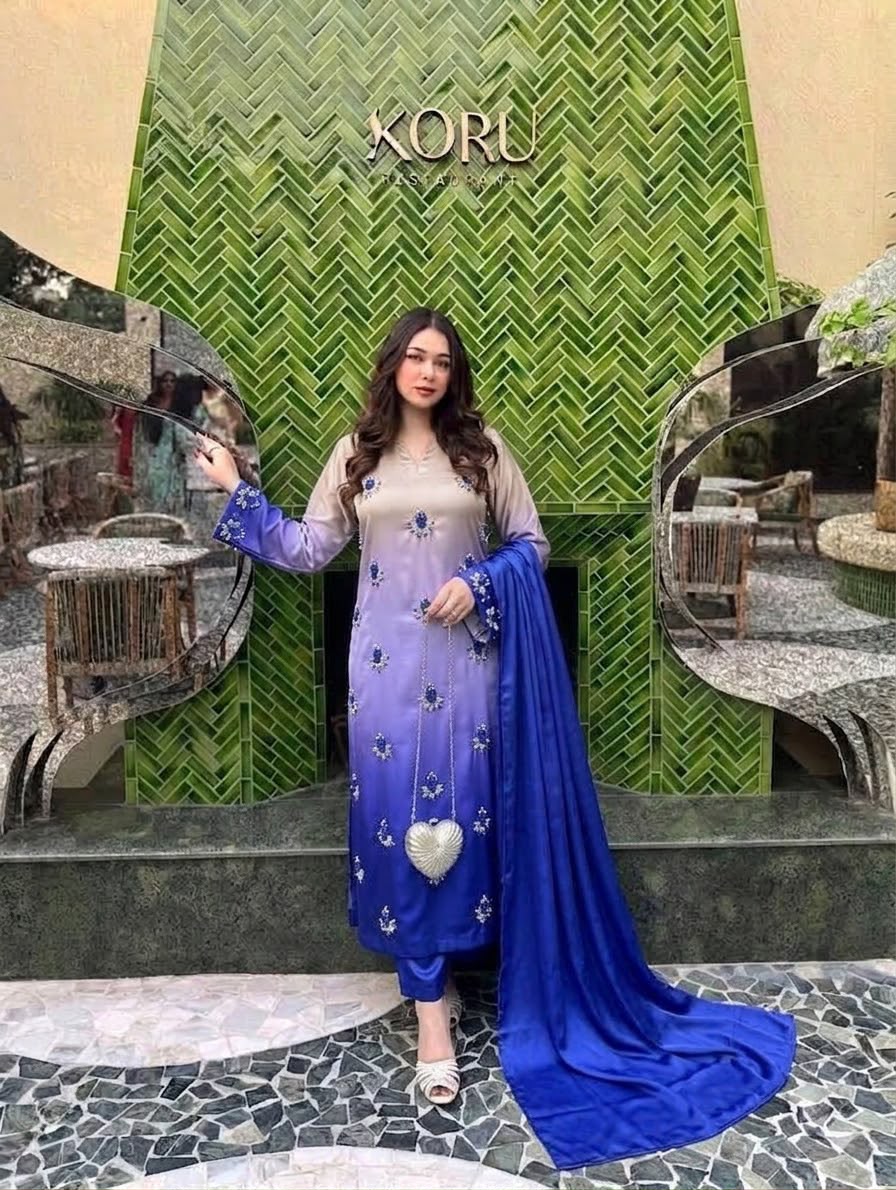 A woman models a traditional South Asian outfit consisting of a purple gradient tunic with blue floral embroidery, matching pants, and a long royal blue dupatta, set against a green herringbone-patterned wall with the brand name KORU visible.