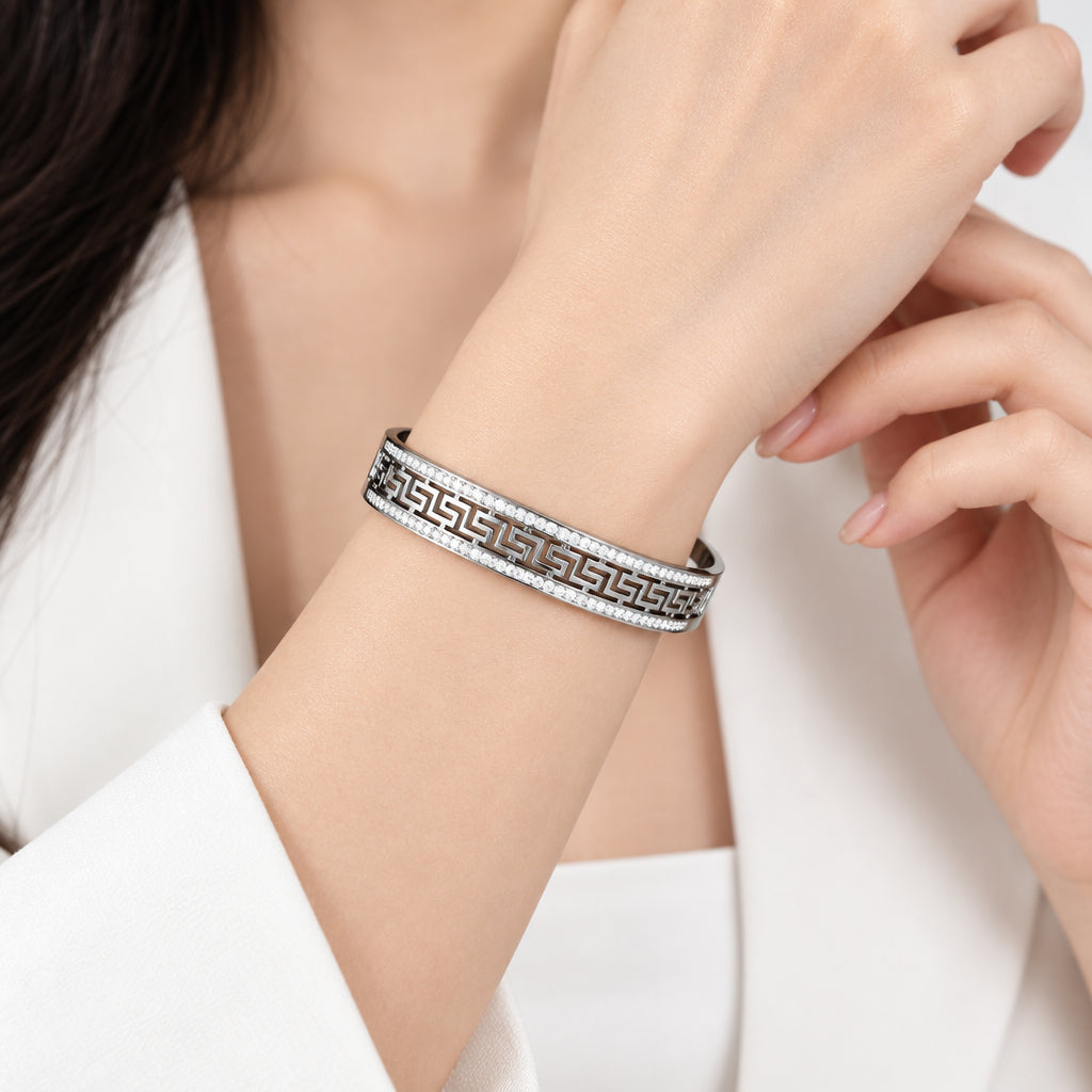 A silver-toned metal bangle bracelet with a Greek key pattern, displayed on a white stone against a soft gray background.