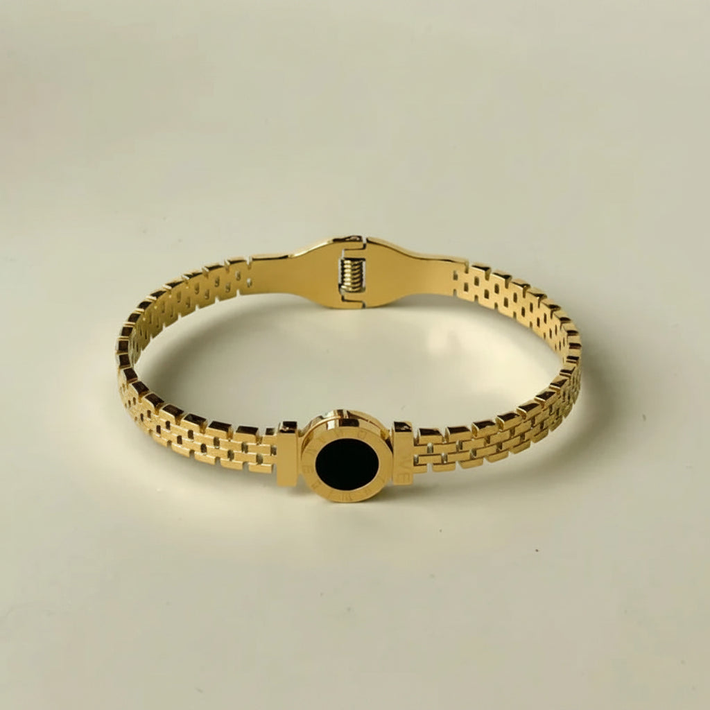 A gold-tone metal link bracelet with a central circular black inlay, displayed on a light beige background.