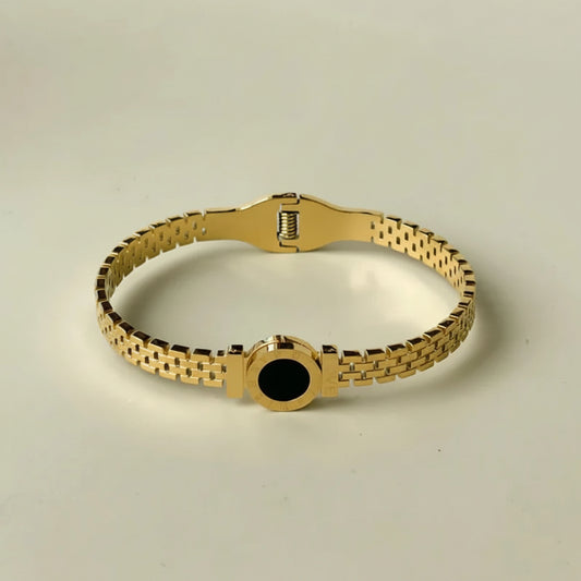 A gold-tone metal link bracelet with a central circular black inlay, displayed on a light beige background.
