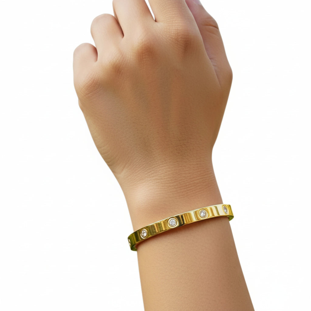 A gold-toned metal bracelet with small clear stones, worn on a person's wrist against a blurred outdoor garden background.