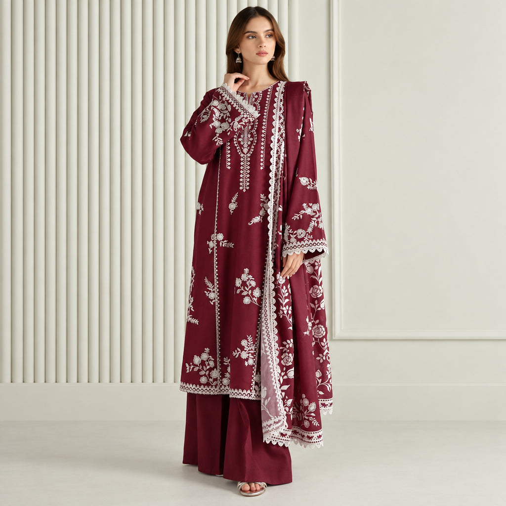 A woman models a three-piece maroon salwar kameez suit with white floral embroidery and lace borders. The set includes a long-sleeved tunic, matching trousers, and a dupatta, shown against a neutral cream-colored background with vertical paneling.