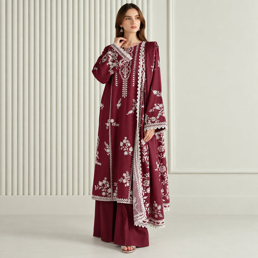 A woman models a three-piece maroon salwar kameez suit with white floral embroidery and lace borders. The set includes a long-sleeved tunic, matching trousers, and a dupatta, shown against a neutral cream-colored background with vertical paneling.