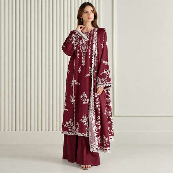 A woman models a three-piece maroon salwar kameez suit with white floral embroidery and lace borders. The set includes a long-sleeved tunic, matching trousers, and a dupatta, shown against a neutral cream-colored background with vertical paneling.