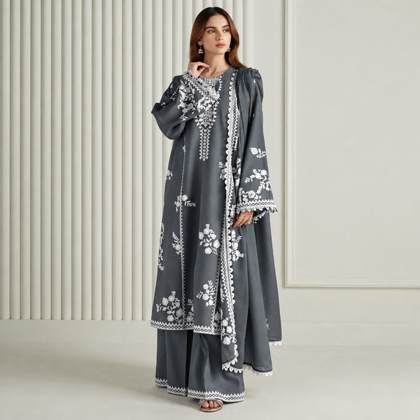 A woman models a dark gray three-piece traditional outfit with white floral embroidery and lace trim, consisting of a long kurta, matching pants, and a draped scarf, set against a neutral cream-colored paneled background.