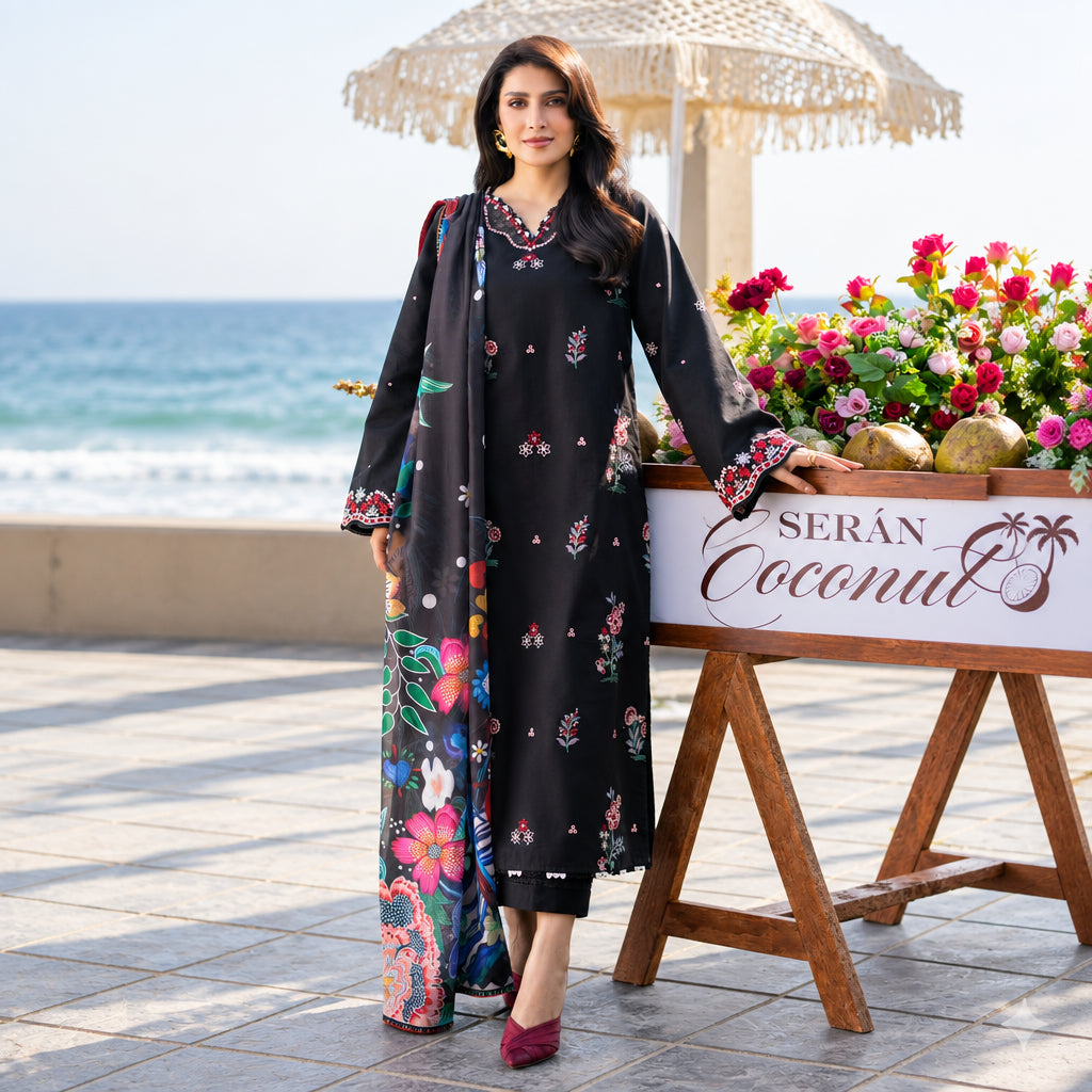 A woman models a black three-piece ethnic outfit with colorful floral prints and embroidered details, standing on a tiled terrace with the ocean and a beach umbrella in the background.