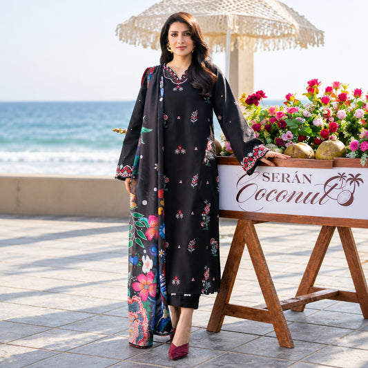 A woman models a black three-piece ethnic outfit with colorful floral prints and embroidered details, standing on a tiled terrace with the ocean and a beach umbrella in the background.