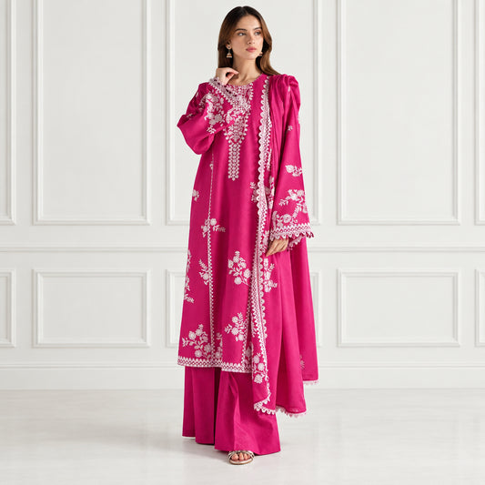 A woman models a bright magenta pink traditional outfit with white floral embroidery and lace details, consisting of a long kurta and matching pants, set against a neutral cream-colored background with vertical paneling.
