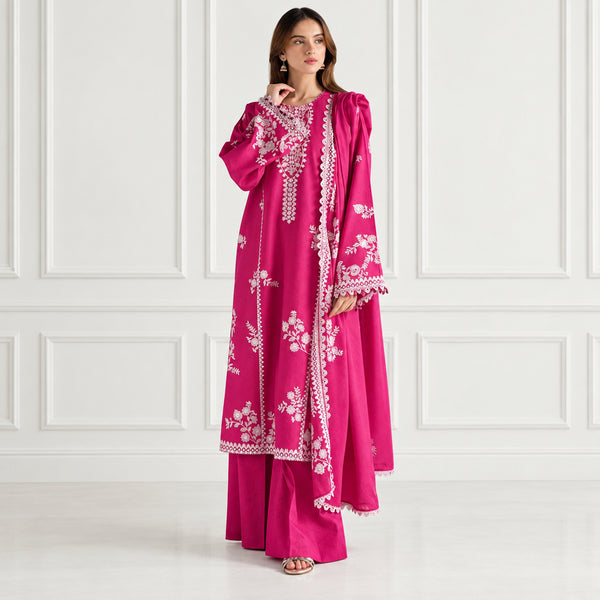 A woman models a bright magenta pink traditional outfit with white floral embroidery and lace details, consisting of a long kurta and matching pants, set against a neutral cream-colored background with vertical paneling.