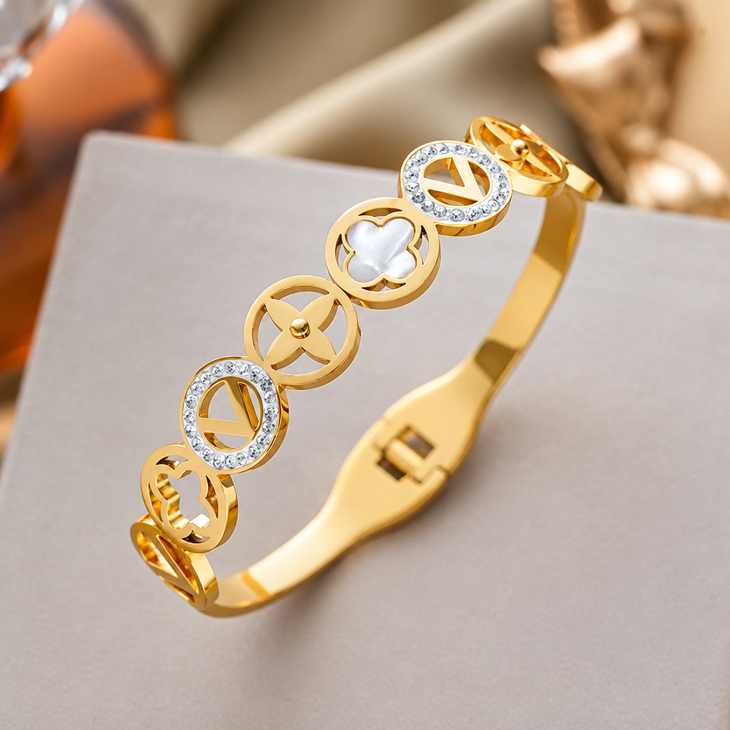 A gold-plated bangle bracelet with circular motifs, including some set with clear crystals and one featuring a white inlay, displayed on a neutral surface.
