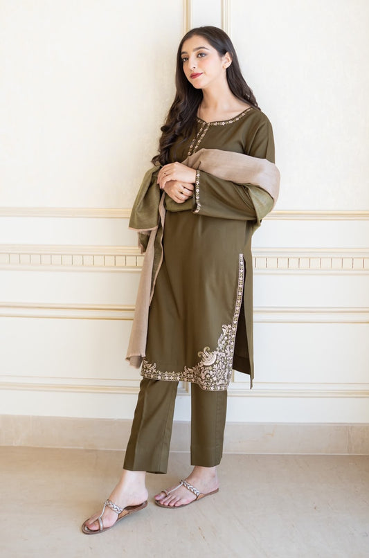 Woman wearing an olive green traditional outfit with a beige scarf against a white wall.