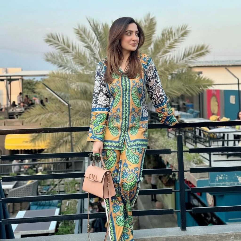 A woman stands on an outdoor balcony wearing a colorful printed two-piece outfit with a yellow base, green and blue paisley motifs, and black-and-white patterned sleeves. She is holding a light pink handbag, with palm trees and a city setting in the background.