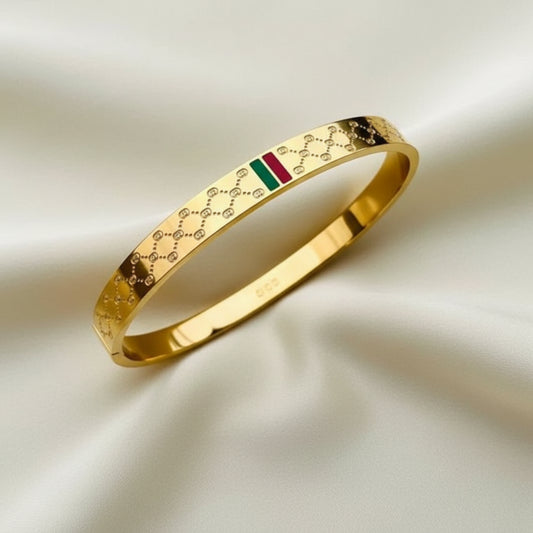 A gold-plated bangle bracelet featuring a textured geometric pattern and a small green and red stripe detail, resting on a soft white fabric background.