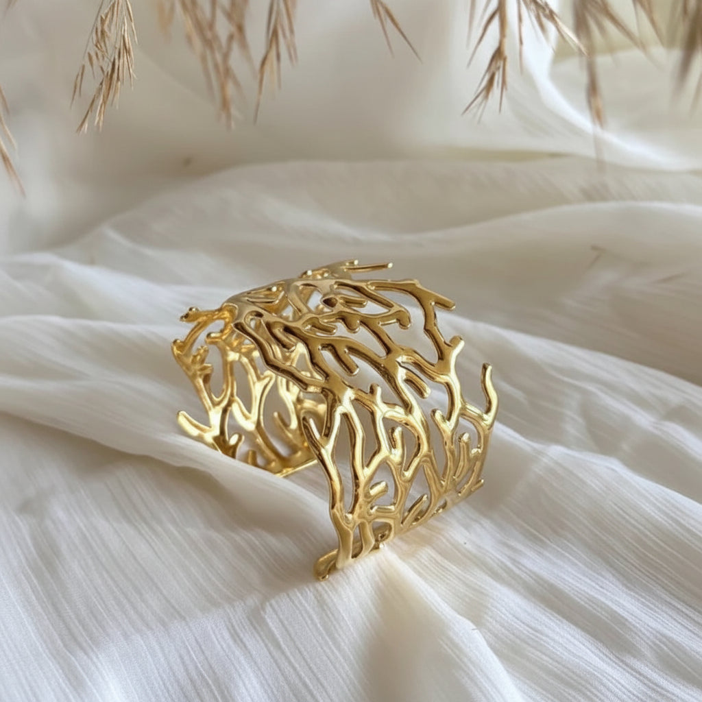 A gold-toned openwork cuff bracelet with a coral or branch-like organic pattern, displayed on draped white fabric with dried grass accents.