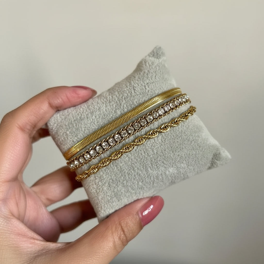 Three gold-toned bracelets displayed on a small gray velvet jewelry cushion, held by a hand with pink manicured nails against a plain light beige background.