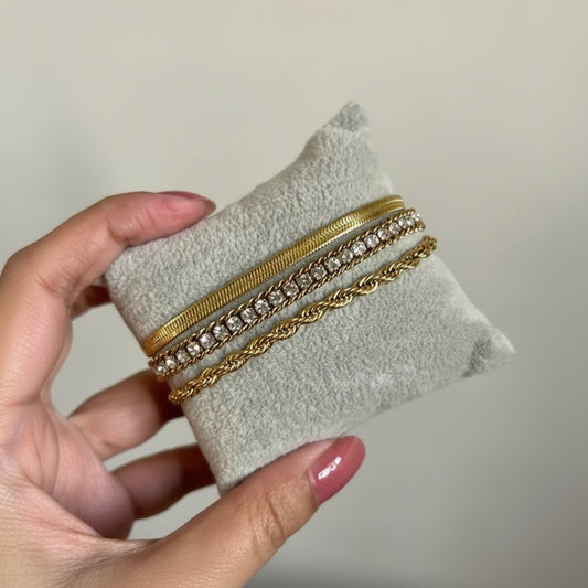 Three gold-toned bracelets displayed on a small gray velvet jewelry cushion, held by a hand with pink manicured nails against a plain light beige background.
