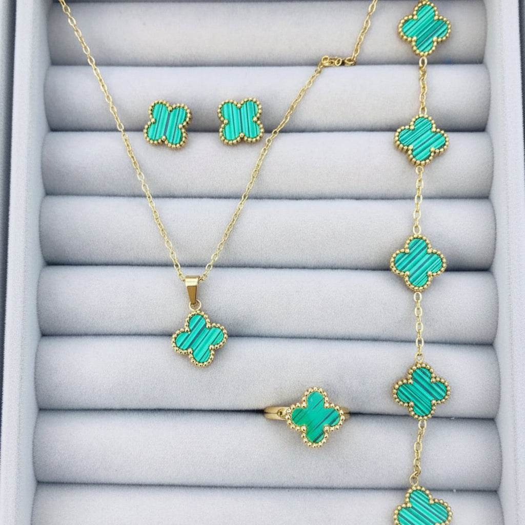 Gold jewelry set with four-leaf clover motifs, including a necklace, ring, and earrings, featuring vibrant turquoise green inlays on a gray display cushion.