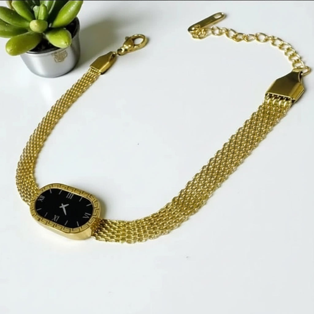 A gold-tone mesh bracelet watch with a black dial and gold-tone case, displayed on a white surface next to a small potted succulent.
