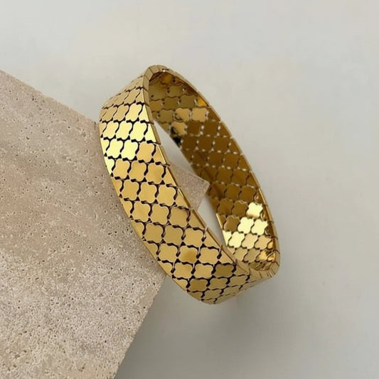 A gold bangle bracelet with a repeating quatrefoil pattern, resting on a textured beige stone block against a light gray background.