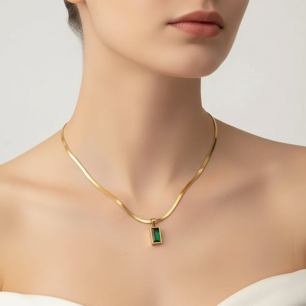 A gold snake chain necklace with a rectangular green pendant, displayed on a draped cream satin fabric background.