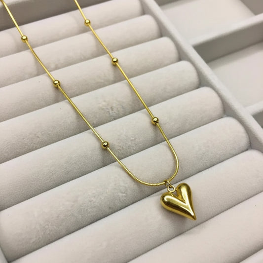 A gold necklace with a small heart-shaped pendant and small bead details on the chain, displayed on a light gray jewelry box.