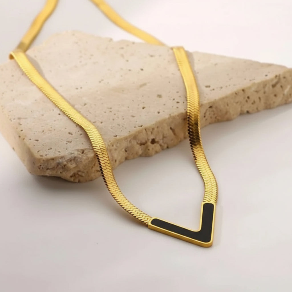 A gold snake chain necklace with a geometric V-shaped pendant featuring a black inlay, displayed on a textured beige stone prop against a light background.
