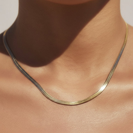A close-up of a delicate gold snake chain necklace worn on a person's neck and upper chest, with warm natural sunlight casting soft shadows.