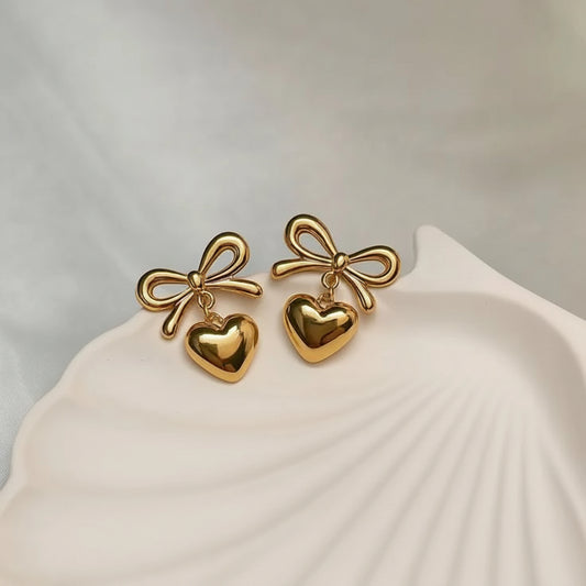 Gold bow and heart earrings on a shell-shaped stand with a neutral background