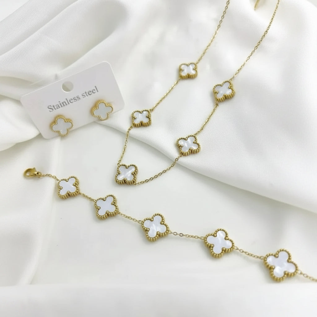 Gold-tone jewelry set featuring a necklace, bracelet, and matching stud earrings with white clover motifs, displayed on a white fabric background with a small card labeled 'Stainless steel'.